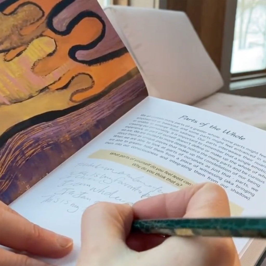 Person holding a pencil and writing in Wisdom of Wholeness: Shadow Work Journal, in the section about parts of the whole, with a colorful painting on the other page.